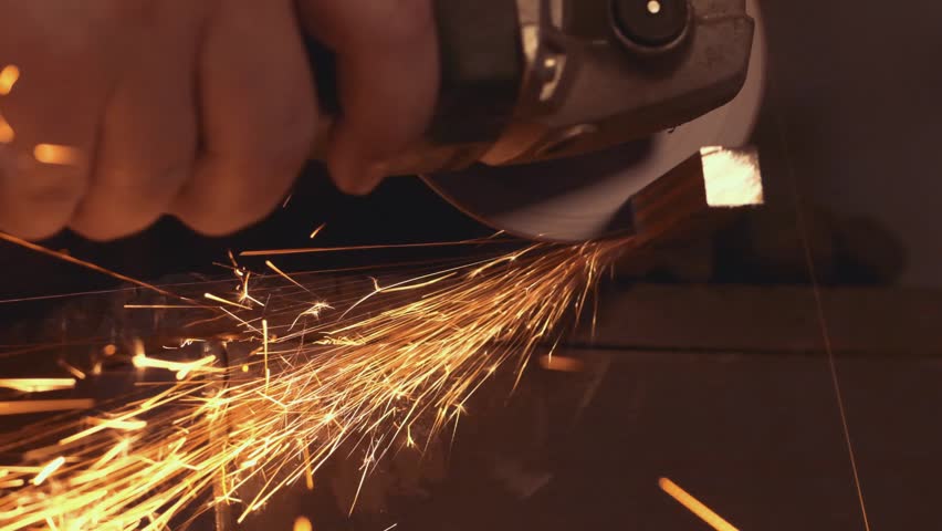 Construction worker cutting metal with angle grinder, generating bright sparks flying against shadowy workshop background, highlighting industrial precision and skilled manual labor