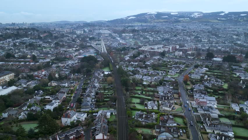 Aerial view of suburban South Dublin with train tracks, modern housing, and distant snowy mountains. Winter urban landscape with residential rooftops and soft overcast skies.