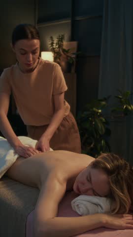 Vertical shot of masseuse performing back massage on woman in softly lit eco-friendly spa room