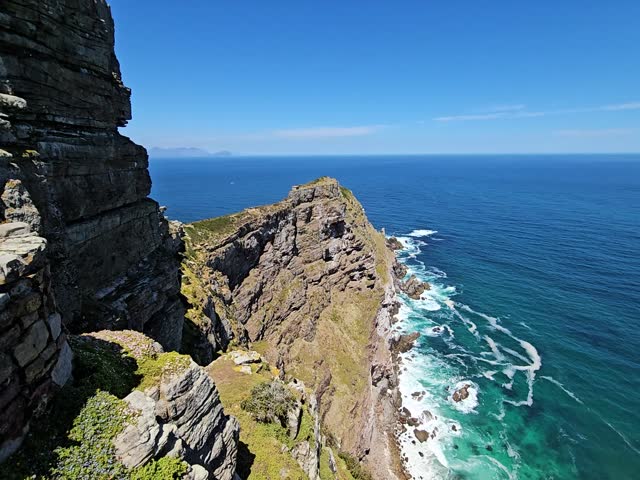 Rocky cliff overlooks the ocean. The water is blue and the sky is clear. There are some rocks on the shoreline in Cape of good hope in South Africa