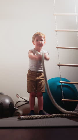 Blond little boy waves battle rope standing near gym equipment. Strong toddler tries effective exercise for strengthening body at home slow motion