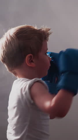 Little boxer in gloves hits punching bag hanging in gym. Blond boy in t-shirt learns boxing basics enjoying professional training slow motion