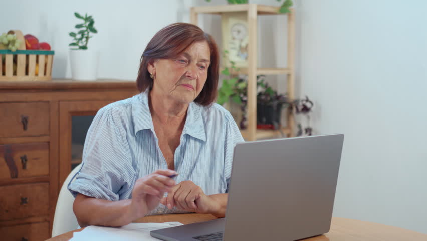 Focused mature woman sitting at table at home looking at laptop and taking notes while freelancing, showing concentration and productivity in remote work environment