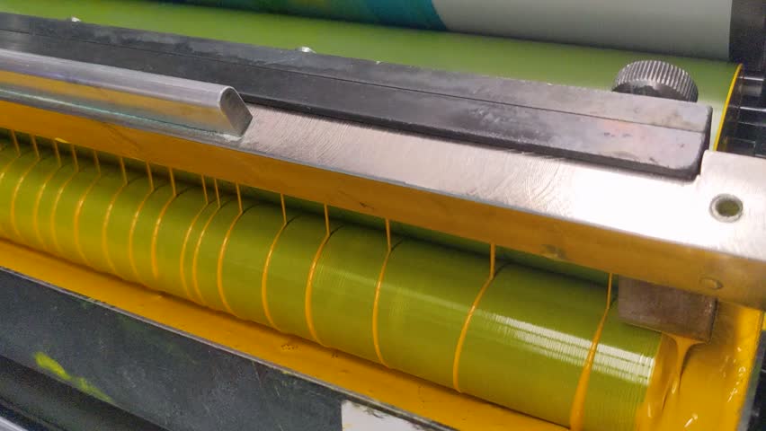 Close-up of an industrial printing press roller applying yellow ink, showing mechanical details and liquid texture