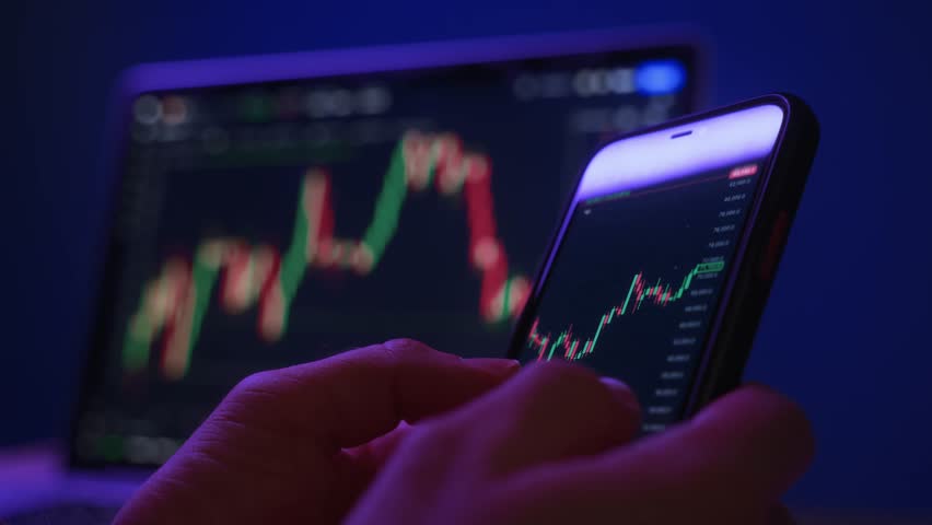 Close-up of hands holding smartphone displaying a stock market candlestick chart, with laptop screen in background. Trader analyzing financial charts on a phone late at night