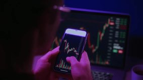 Over the shoulder view of a trader closely examining cryptocurrency charts on a smartphone via a trading app, with stock market data displayed on a laptop in the background - Powered by Shutterstock - Get 15% off with code: PIKWIZARD15