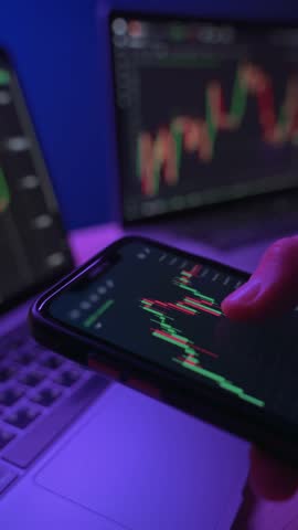 Close up real-time market chart with price indicators on smartphone held by trader or crypto investor in dark neon lit workspace. Businessman monitoring crypto via trading app. Vertical video