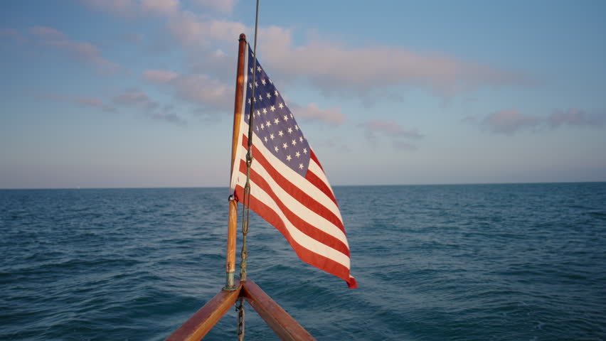 An American flag waves gently in the soft evening breeze on the stern of a beautifully crafted sailing ship, as the magnificent sun sets over the tranquil and picturesque waters of Key West, Florida