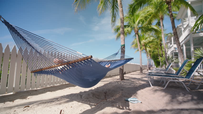 A woman relaxes in a hammock at a beautiful Key West hotel, surrounded by palm trees and a sandy beach, enjoying peaceful moments during her vacation, soaking up the sun and feeling rejuvenated