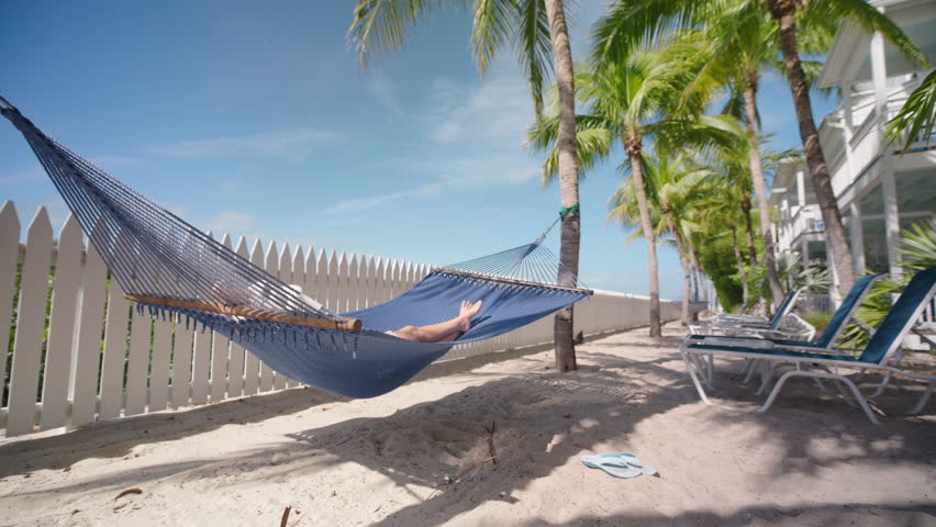 A woman relaxes in a hammock at a beautiful Key West hotel, surrounded by palm trees and a sandy beach, enjoying peaceful moments during her vacation, soaking up the sun and feeling rejuvenated