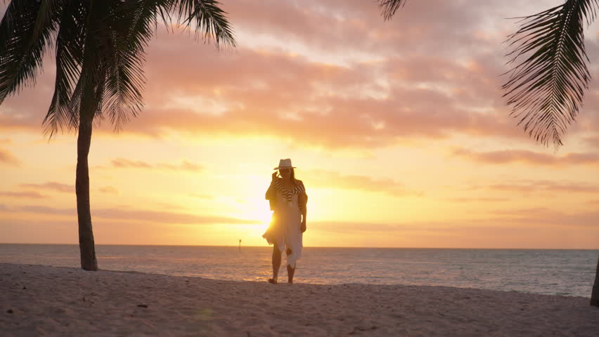 A woman enjoys a peaceful morning walk on the beach at sunrise in Key West, Florida, soaking in the tropical beauty, golden light, and relaxing vibes of a calm seaside moment in slow motion.