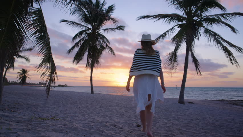A woman strolls along the quiet beach at sunrise in Key West, Florida, surrounded by gently swaying palm trees and the sound of waves, capturing a moment of serenity and tropical beauty. 