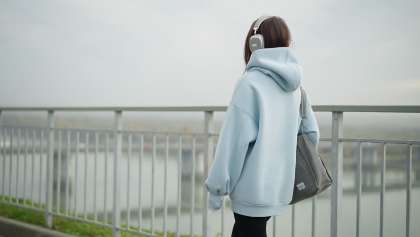 Side view of beautiful girl in casual hoodie walking with bag near iron rail, blurred view of bridge and river in background, creating a calm, urban atmosphere perfect for lifestyle content