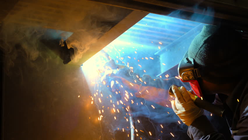 Welder at work, wearing protective gear, welding helmet and heavy-duty cloth. Bright blue and orange sparks are flying as the welding torch fuses metal together