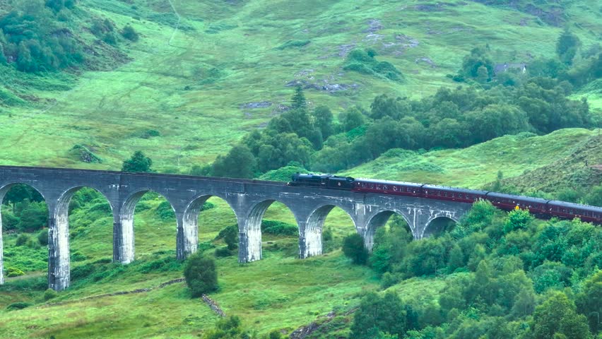 Hogwarts Express train crossing a bridge. the hills are green and the train blows steam as it finishes crossing