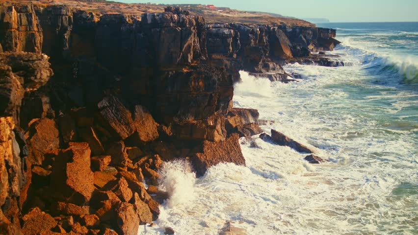 Slow motion video of the huge ocean waves  crashing onto the rocky shore of coastline in Portugal. Beautiful big ocean waves are rolling onto shore and crushing on rocks. Oceanic tide during sunset.