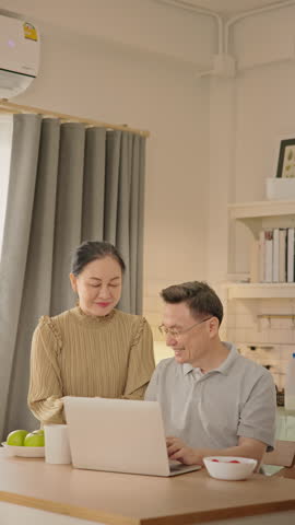 Senior couple enjoying peaceful morning together at home. Elderly Asian man and woman sit together smiling and talking with laptop and breakfast on the table. healthy aging, retired lifestyle.