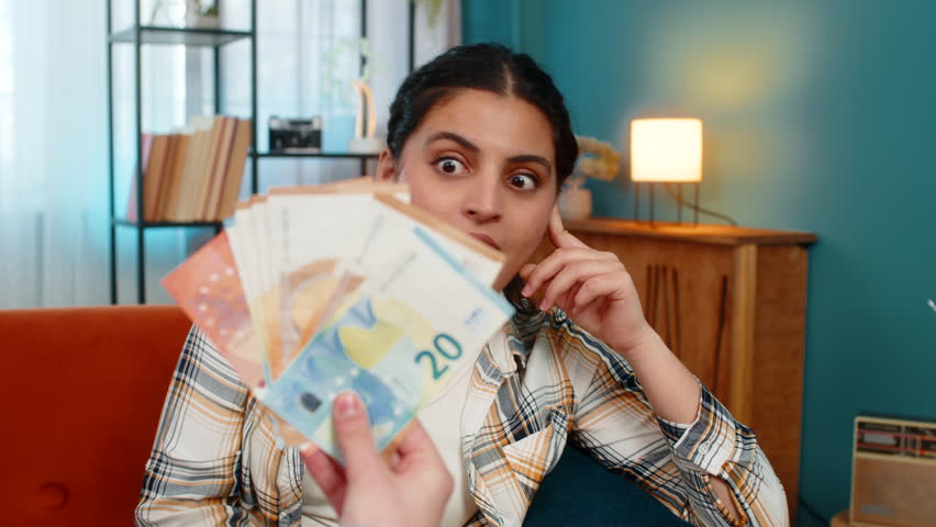 Indian happy woman experiencing sudden happiness, joyfully celebrating on sofa receiving plenty of cash euros. Arabian girl sincerely delighted, smiling widely, enthusiastically showing victory win