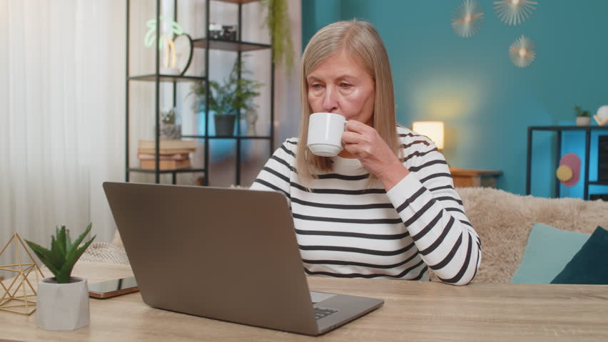 Mature woman working from home on laptop, writing report or replying to emails. Grandmother sitting at table enjoying coffee while managing daily tasks with concentration and dedication at home office