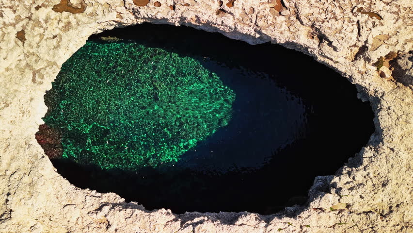 Turquoise waters are visible through a large rock opening on the coastline of Malta. The area is surrounded by rugged terrain and showcases natural beauty.