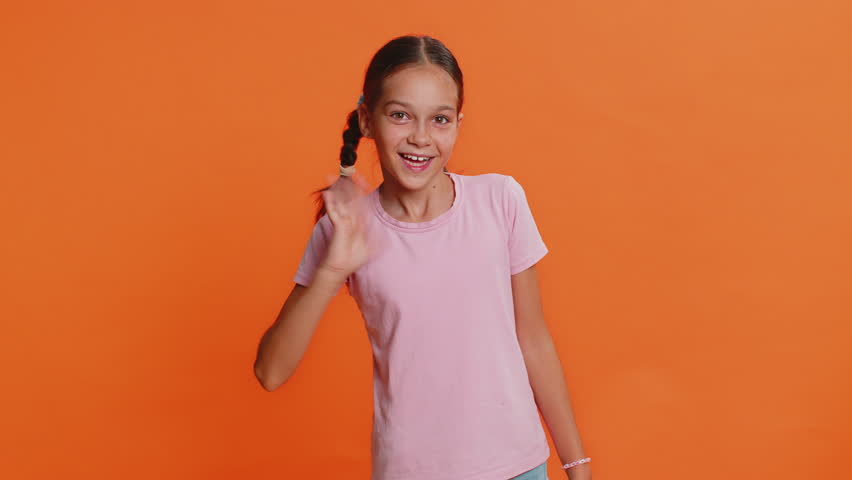 Young cute school girl 10 years old smiling friendly at camera and waving hands gesturing hello or goodbye, welcoming with hospitable expression. Preteen female child kid isolated on orange background