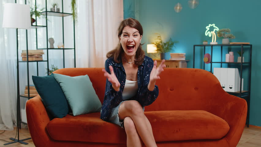 Portrait of happy young woman female with brown hair watching TV celebrating success dancing sitting on sofa couch in living room at home. Excited adult girl in casual clothes winning match at home.