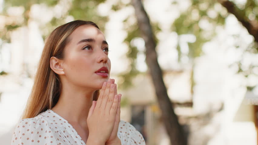 Portrait of young woman praying with closed eyes to god asking for blessing, help, forgiveness outdoors. Pretty girl clasping hands wishing luck in sunny urban city street. Town religion lifestyles.