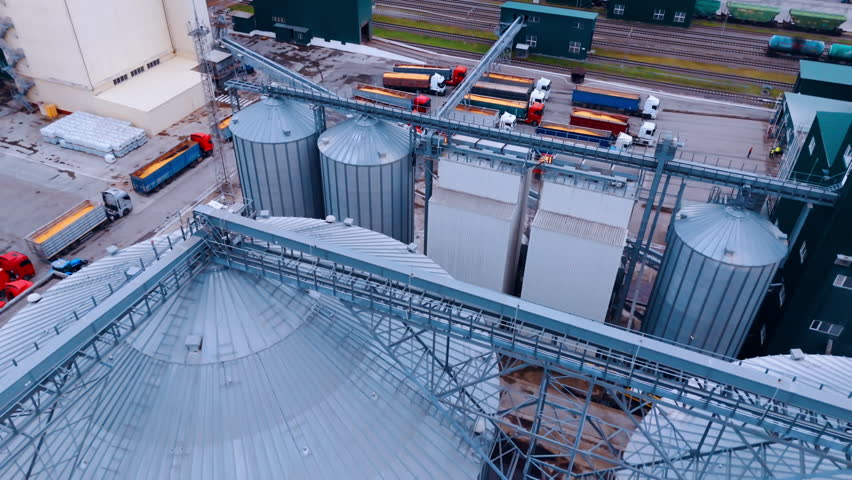 Flight above the steel tanks of granary. Loaded trucks full of grain stand at the territory of the plant at backdrop.