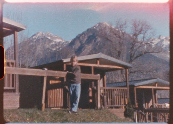 Vintage, slightly shaky footage shows a blond girl standing in front of a wooden fence in a mountain campsite, with snowy peaks in the background