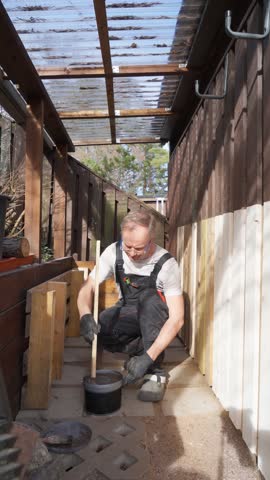 Man painting wooden the old barn siding outdoors during renovation. Wearing work overalls and gloves, he applies paint to wood planks. DIY, craftsmanship, home improvement, and exterior maintenance.