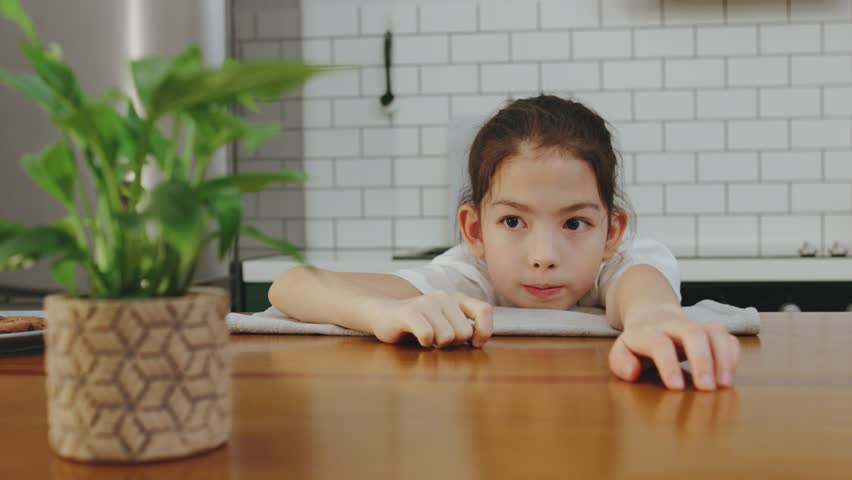 Young child with focused expression reaches out over a wooden table in a bright kitchen surrounded by modern decor and plants during daytime