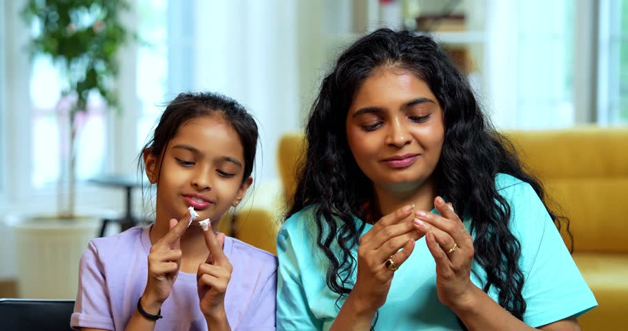 Indian Asian young woman and cute little girl happily and playfully applying face cream on cheeks while smiling and looking in the mirror together at home, enjoying fun skincare beauty time indoors