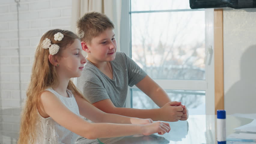 Hand view giving white paper to two kids sitting on chair boy on gray top girl on white gown indoor, focus on paper and children in bright room with window background