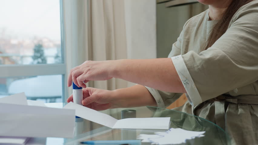 Hand view of woman in gray gown applying glue on paper craft, focus on hands using glue stick for crafting in cozy home setting with snow outside window in background