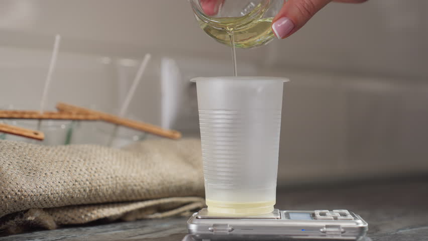Close-up hand pouring baking liquid from glass cup into measuring cup on digital scale with soft cotton cloth and wooden wicks visible in background kitchen setting