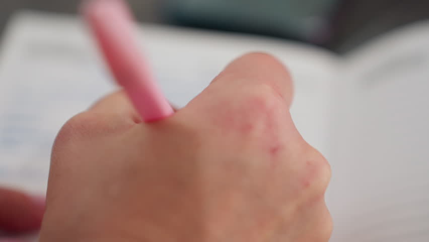Close up hand view of white person writing with pink pen on paper with soft blur surroundings showing concentration and gentle motion, with natural lighting in background