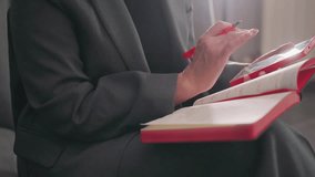 Businesswoman writing in red notebook with pen, focused on work, modern office environment, gray suit, sitting relaxed, productivity, decision-making, corporate workspace, concentration - Powered by Shutterstock - Get 15% off with code: PIKWIZARD15