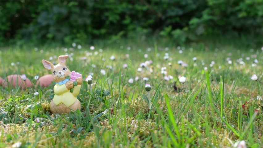 Five toy rabbits lined up on green grass with daisies in the background. The camera smoothly pans from right to left, capturing the charming scene in natural daylight. Easter decoration theme.