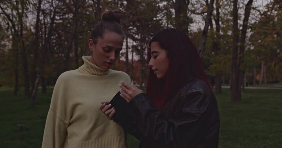 Two young women are looking at a wallet in a park and reacting with surprise and happiness to what they find inside.