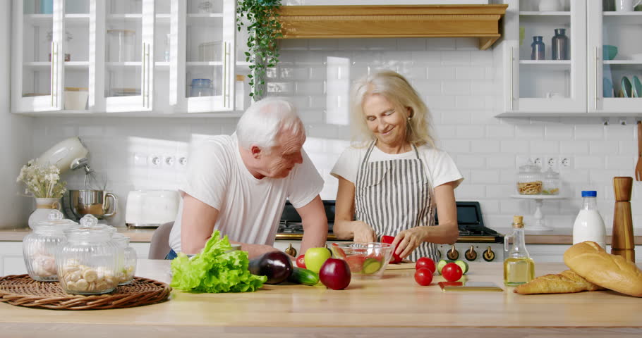 Aged woman playing with slices of paprika when cooking salad, joke for husband. Elderly married couple cutting vegetables for dinner or lunch, good mood and positivity, retirees having fun and joy