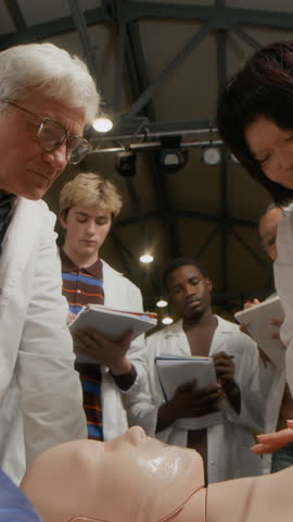 Vertical shot of of Asian teenage girl pushing manikins chest training practicing cardiopulmonary resuscitation while male professor providing guidance in group class at college