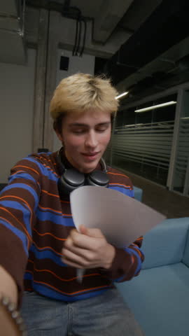 Vertical shot of excited teenage boy in casual clothes talking on video call using phone showing sheet of paper with test results while sitting in common area of campus