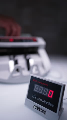 Red numbers increasing on the bill counter display. One hundred dollar bills are calculated in cash counting machine at blurred backdrop. Vertical video.