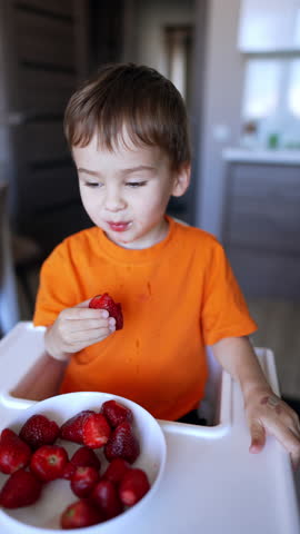 Lovely smiling baby boy sits at high chair eating strawberry. Healthy eating habits and vitamins consumption. Vertical video.
