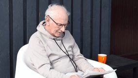 Elderly man leisurely reading newspaper, sitting in white chair on sunny patio, holding orange mug with steaming drink - Powered by Shutterstock - Get 15% off with code: PIKWIZARD15