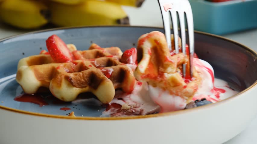 A fork pierces a piece of delicate waffle and dips it into ice cream with strawberry syrup while eating. delicious belgian waffles with vanilla ice cream and fresh strawberries and blueberries