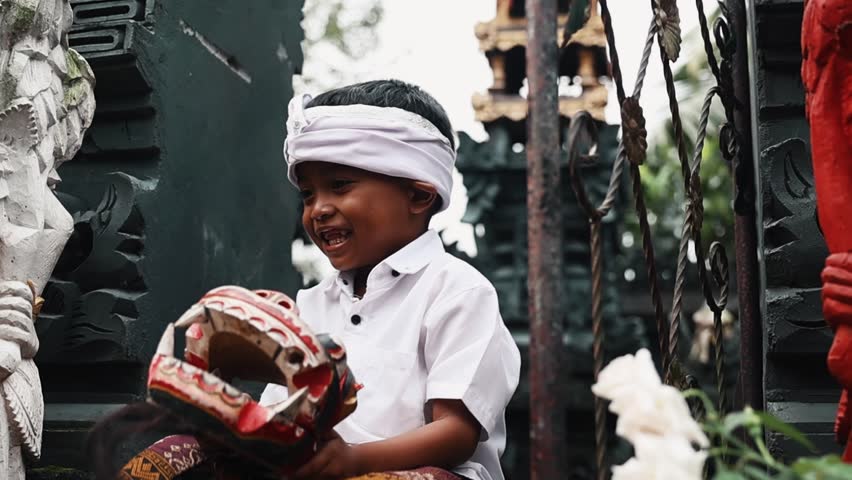 Indonesian boy in traditional Bali attire shaking mask near temple