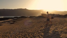 LENS FLARE: Male surfer runs to the beach with wooden surfboard tucked under his arm in golden light. He stops before heading into water to check breaking waves at iconic surf spot on Lanzarote island - Powered by Shutterstock - Get 15% off with code: PIKWIZARD15