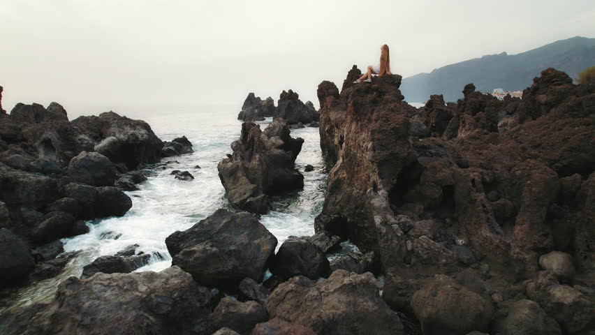 Person sits atop volcanic rocks, ocean waves crash gently against the rugged shore. Charco del Diablo in Tenerife Canary Islands Spain.