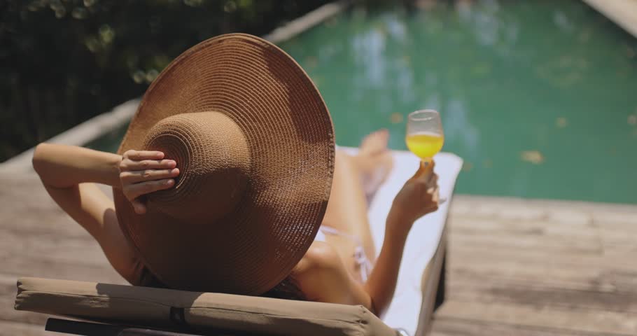 A stylish woman in a wide-brimmed sun hat lounges on a poolside chair, holding a refreshing cocktail. A perfect summer scene for vacation, relaxation, and luxury resort concepts. 